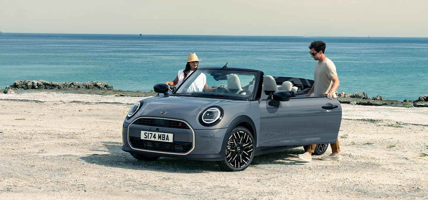 Vista frontal izquierda del MINI Cooper Cabrio en Copper Grey en una playa con dos personas al lado.