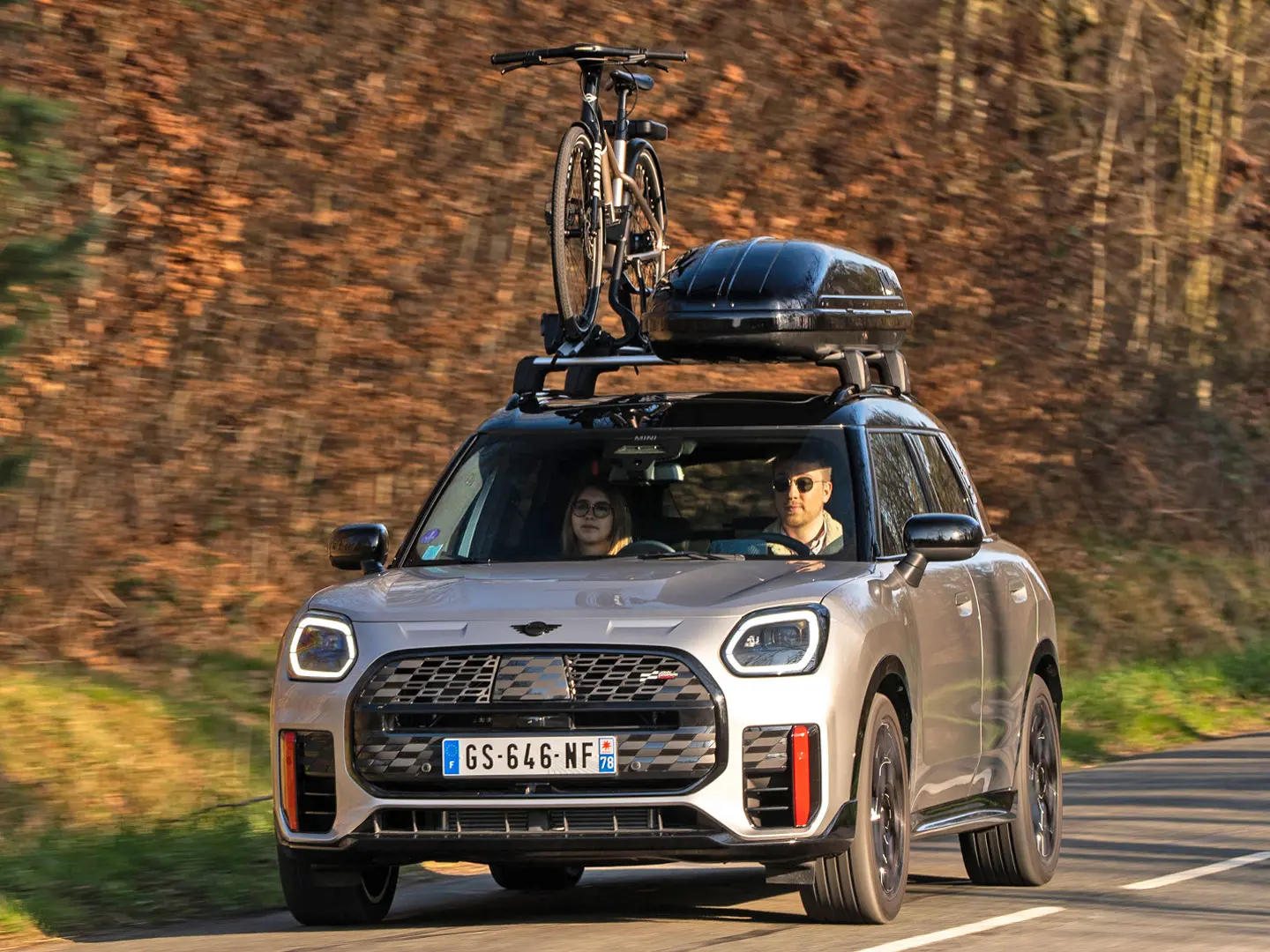 MINI Countryman Cooper en Silver en una carretera de montaña con portaequipajes de techo MINI, caja de techo MINI, portabicicletas de techo MINI con una bicicleta montada y alerón MINI John Cooper Works (vista frontal).