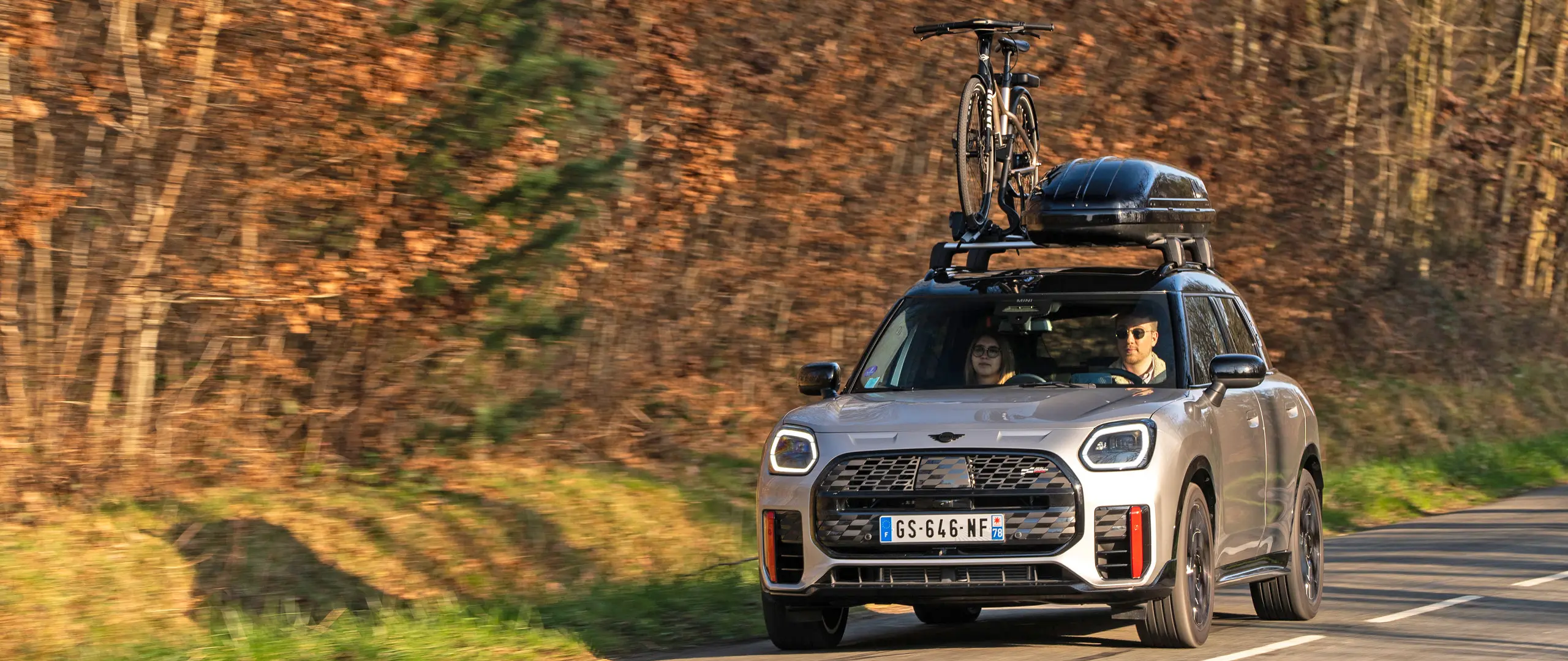 MINI Countryman Cooper en Silver en una carretera de montaña con portaequipajes de techo MINI, caja de techo MINI, portabicicletas de techo MINI con una bicicleta montada y alerón MINI John Cooper Works (vista frontal).