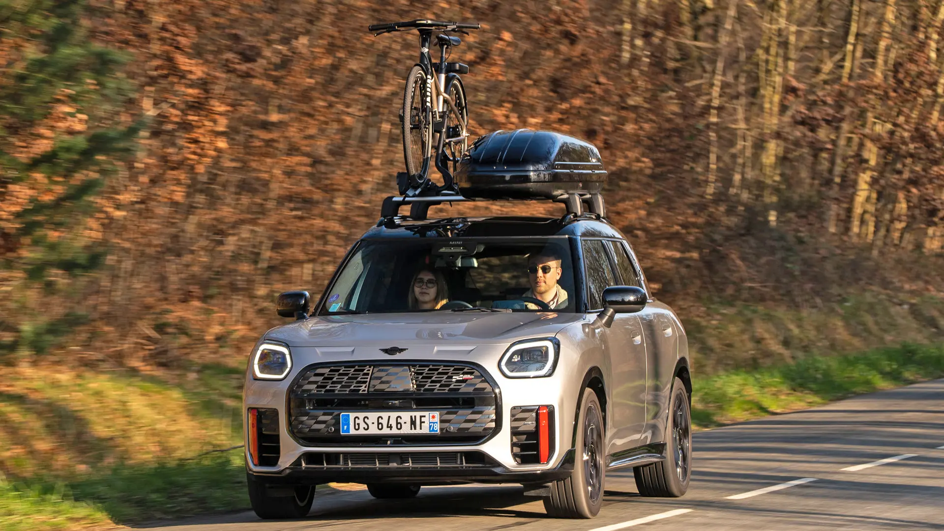 MINI Countryman Cooper en Silver en una carretera de montaña con portaequipajes de techo MINI, caja de techo MINI, portabicicletas de techo MINI con una bicicleta montada y alerón MINI John Cooper Works (vista frontal).