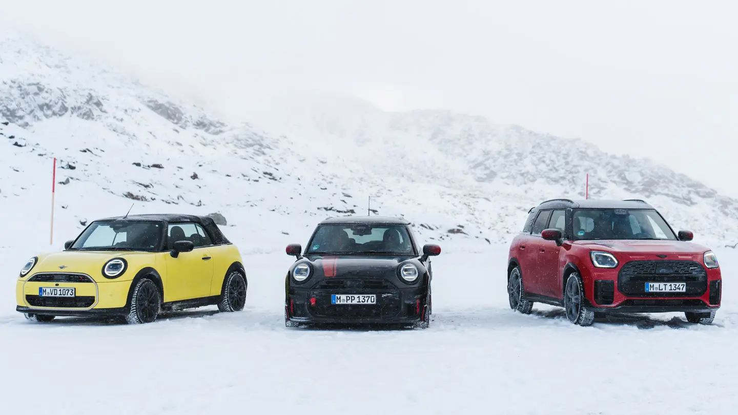 El MINI Cooper descapotable en Sunny Side Yellow, el MINI John Cooper Works en Midnight Black II y el MINI Countryman Cooper en Chili Red parados en una carretera montañosa nevada (vista frontal).