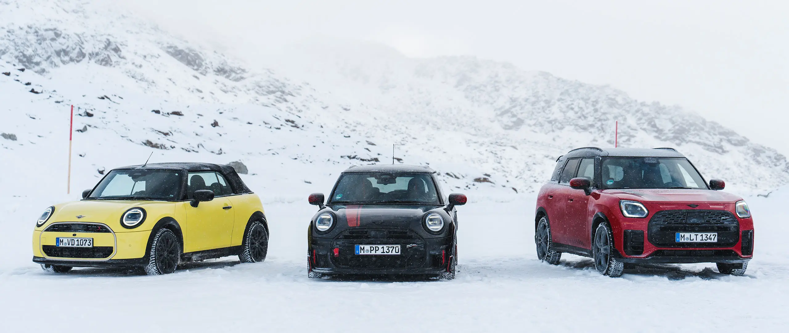 El MINI Cooper descapotable en Sunny Side Yellow, el MINI John Cooper Works en Midnight Black II y el MINI Countryman Cooper en Chili Red parados en una carretera montañosa nevada (vista frontal).