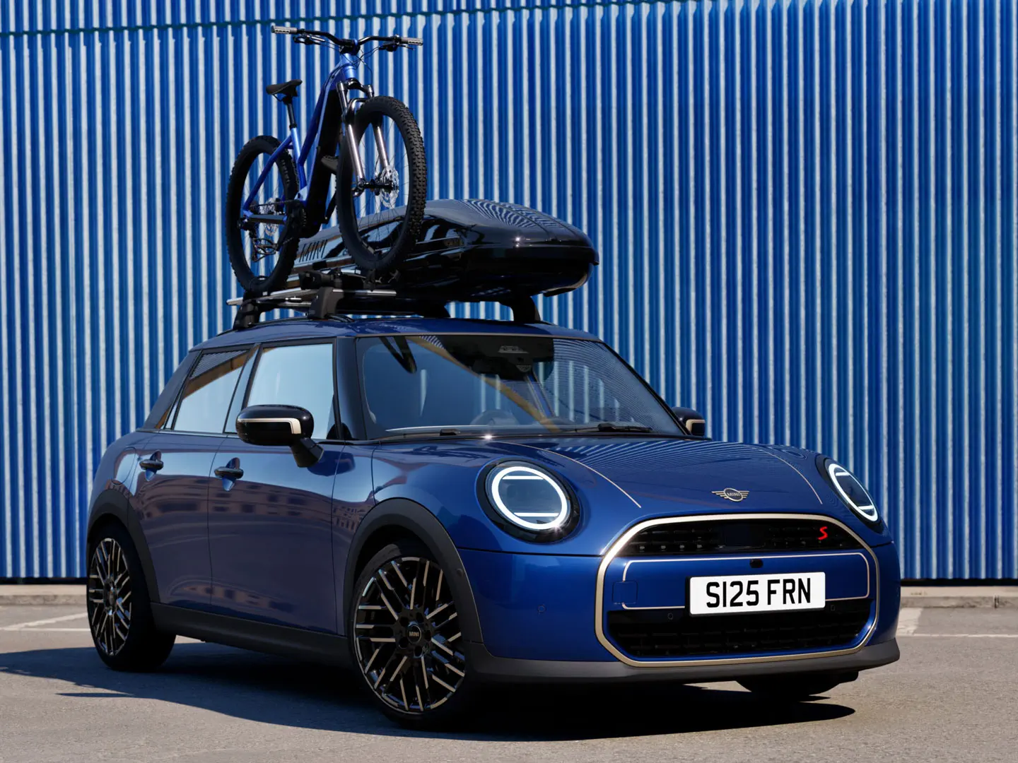 Vista frontal del MINI Aceman en Indigo Sunset Blue con un soporte para bicicletas MINI cargado con una bicicleta de montaña y una caja de techo MINI 320.