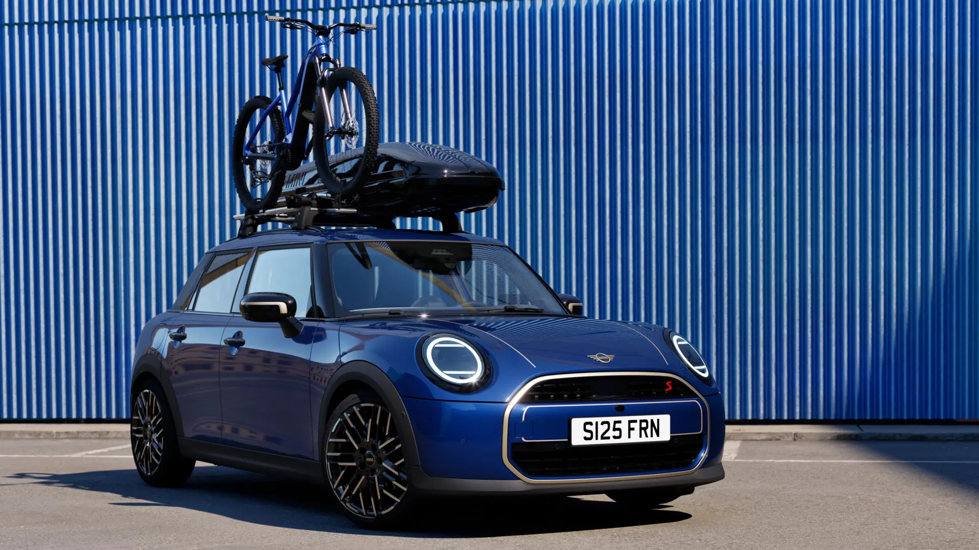 Vista frontal del MINI Aceman en Indigo Sunset Blue con un soporte para bicicletas MINI cargado con una bicicleta de montaña y una caja de techo MINI 320.