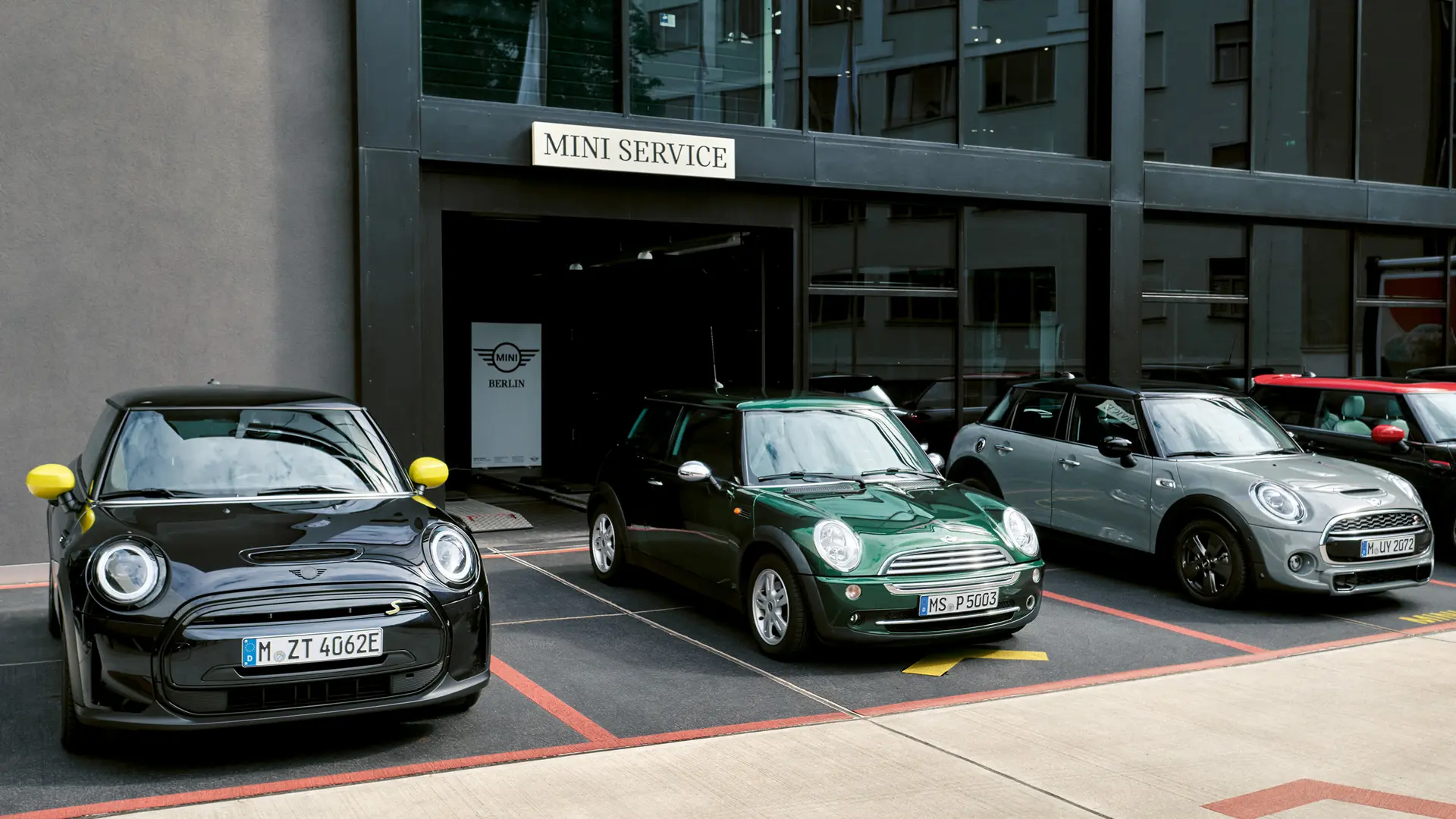 Un MINI Cooper Electric S en Black estático en el exterior de un MINI Centre junto a un MINI Cooper 3 Puertas en Green, un MINI 5 Puertas en Silver y un MINI JCW Black, vista frontal. 