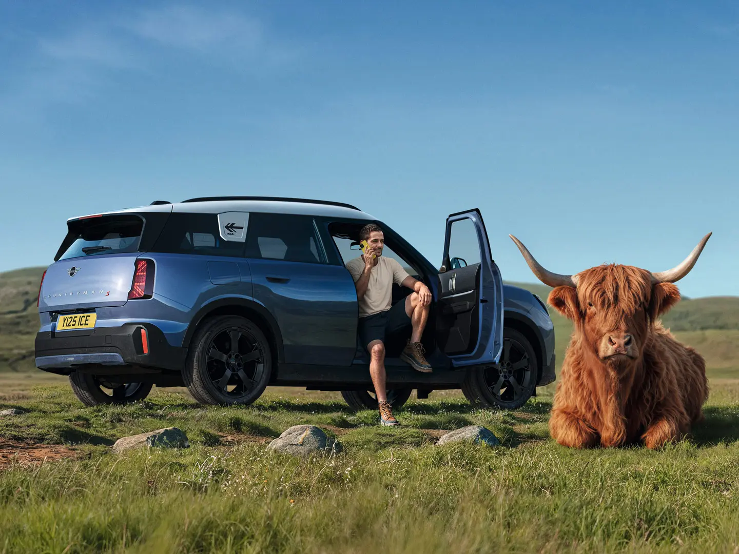 Un hombre está junto a su MINI Countryman S hablando con la asistencia en carretera MINI a través de la MINI App con una vaca de las tierras altas tumbada en las proximidades.