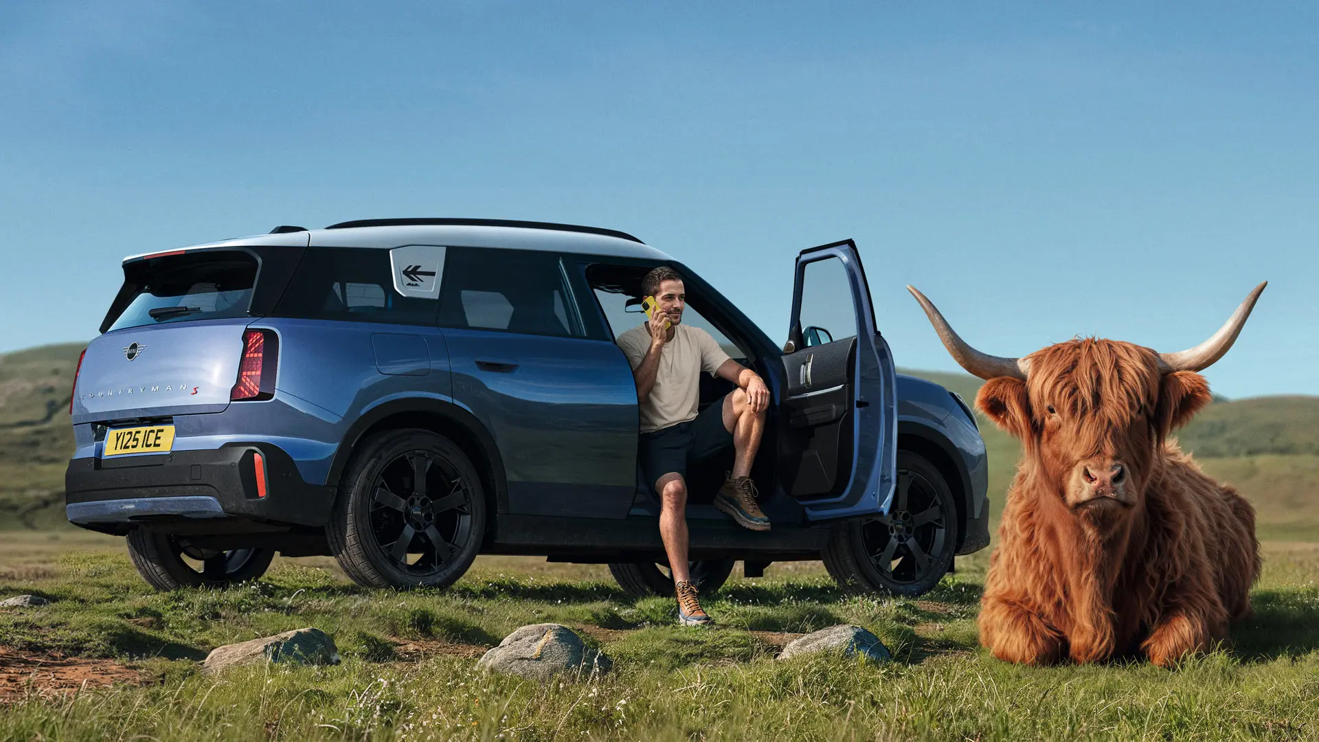 Un hombre está junto a su MINI Countryman S hablando con la asistencia en carretera MINI a través de la MINI App con una vaca de las tierras altas tumbada en las proximidades.