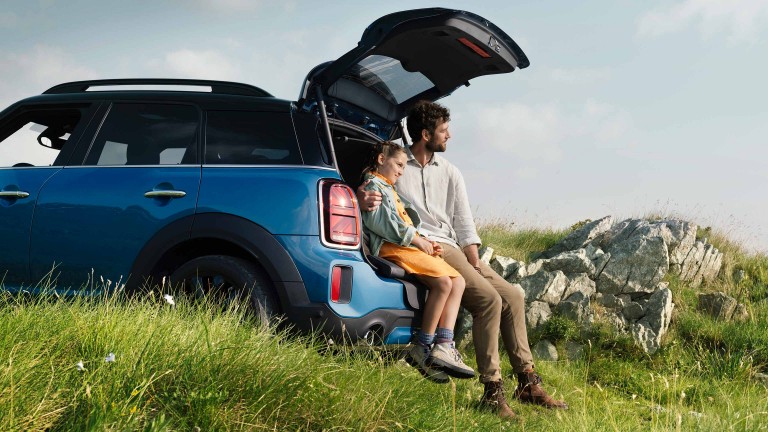 Seitenansicht des MINI Highlands Edition mit Mann und Frau auf der Picknickbank sitzend auf der Ladekante in der Natur